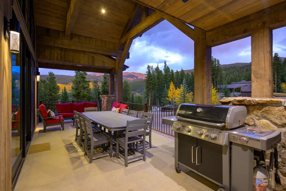 A spacious patio area is furnished with a dining table, grill, and comfortable seating, all oriented to capture sweeping mountain views. The scene evokes the enjoyment of outdoor dining, barbecuing, and relaxed socializing against a dramatic natural backdrop.