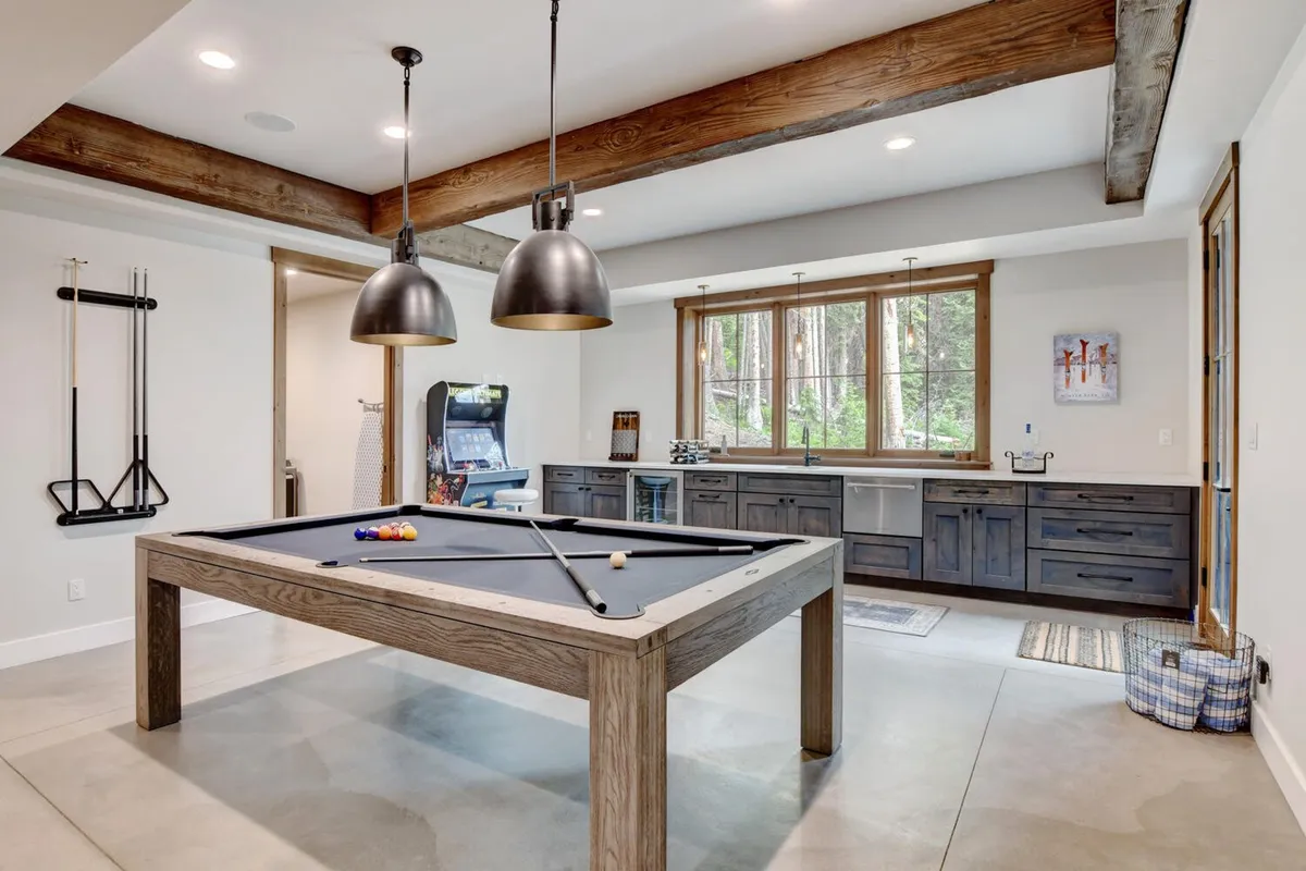 A bright, inviting game lounge featuring a sleek pool table, generous windows with outdoor views, and a kitchenette area in the corner, perfect for prepping snacks and drinks. The room radiates casual fun and sets the stage for group activities.