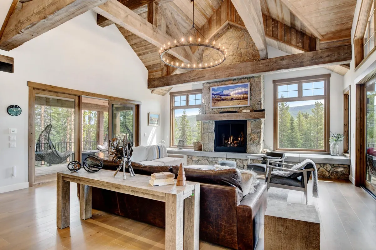 A spacious living room area featuring a grand stone fireplace, comfortable seating, rustic wooden beams, and expansive windows that reveal sweeping mountain views outside. The atmosphere is one of warmth and connection, ideal for group gatherings or relaxing after a day of adventure.