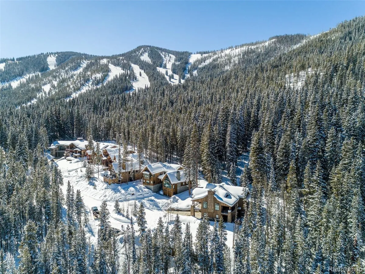 A cluster of snow-covered cabins nestled among dense pine trees, with ski slopes visible in the background. The landscape is tranquil, evoking the quiet beauty of a winter mountain retreat, and hints at direct access to skiing right from the door.