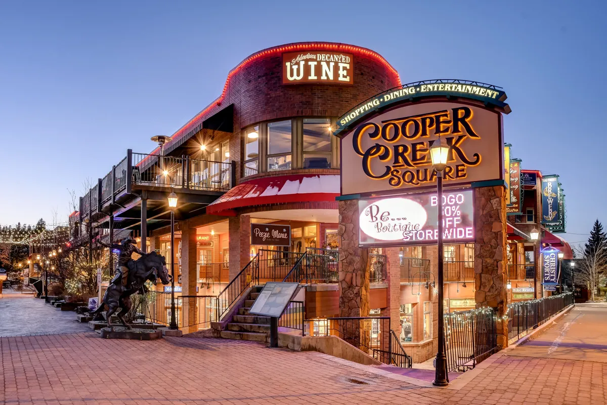 The Cooper Creek Square sign is visible alongside a wine shop sign, an outdoor seating area, and a horse sculpture. This bustling exterior scene highlights shopping, dining, and arts in downtown Winter Park, with a lively and festive air.