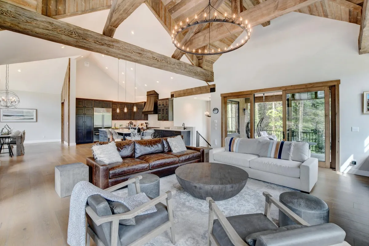 An open-concept living area with a large leather sofa, coffee table, exposed wooden beams, and a kitchen area—all framed by generous windows letting in natural light and views of the outdoors. The space is perfect for socializing or informal team-building activities.