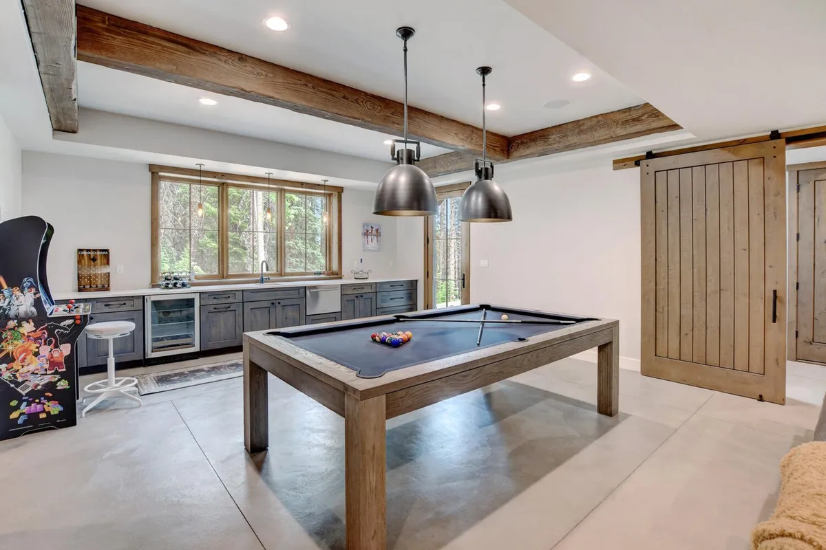 This image captures a vibrant game room with a pool table as the centerpiece, an arcade machine standing against the wall, a kitchenette for snacks or drinks, and large windows letting in natural light. The setup encourages friendly competition and socializing, embodying the spirit of a multi-use entertainment space.