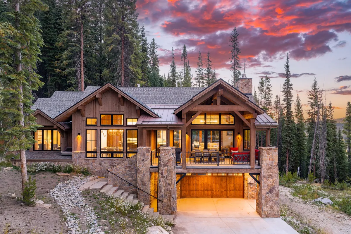 The hero image showcases the luxurious exterior of a mountain chalet surrounded by towering trees, with expansive windows reflecting a vibrant sunset sky. The spacious porch invites guests to unwind while soaking in breathtaking mountain views, and the yard is perfectly set up for outdoor dining and family barbecues.