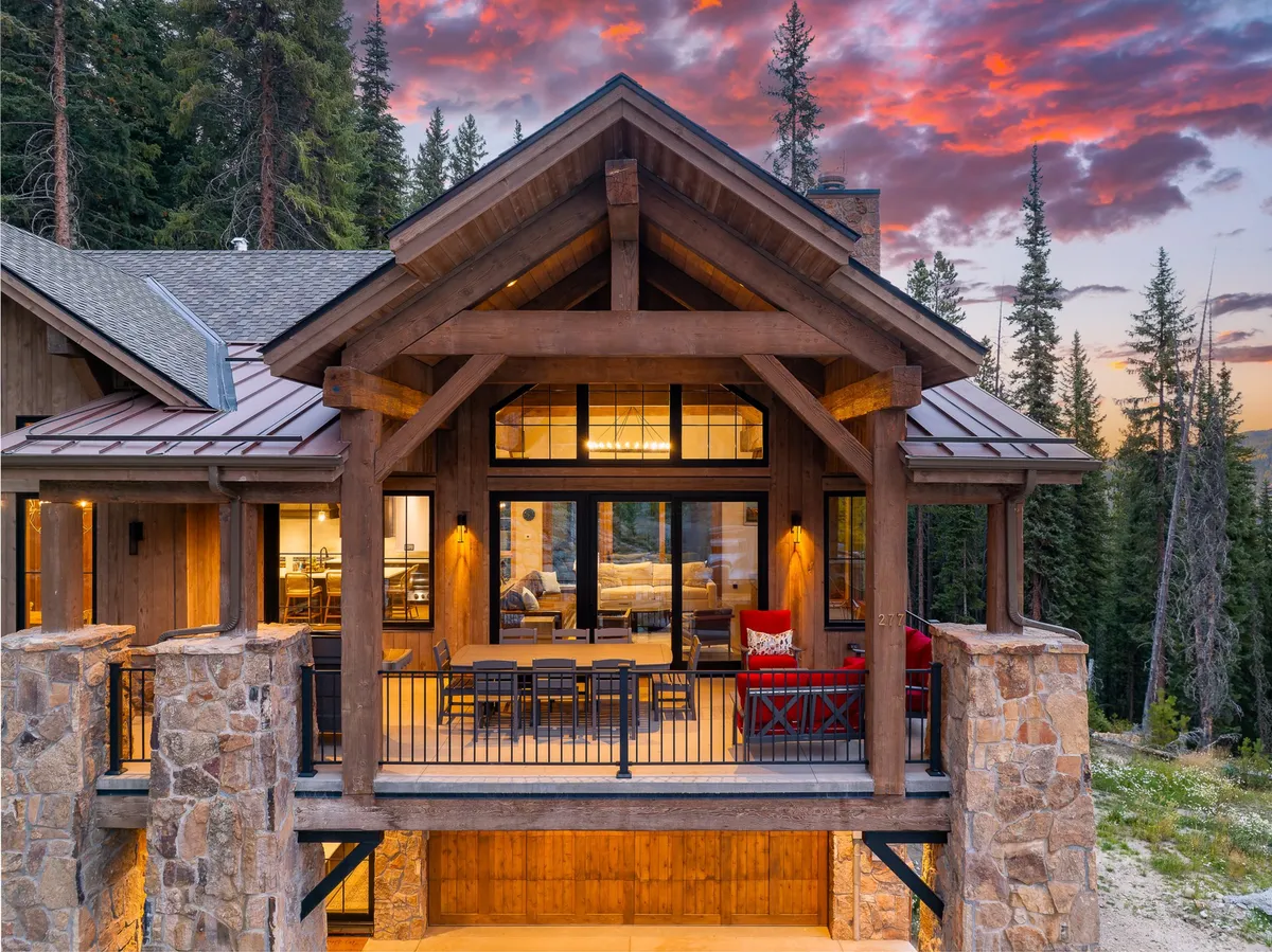 A welcoming exterior view of a chalet-style home featuring large wooden beams, expansive glass windows, stone pillars, and an inviting outdoor seating area. The setting glows with the warmth of sunset, suggesting a cozy retreat after a day on the slopes.