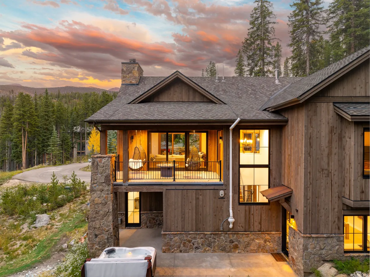 This image showcases the chalet's wooden exterior nestled within a lush forest setting. Large windows invite natural light, and a balcony offers a vantage point for enjoying the views. The scene is set during sunset, with a warm sky and guests relaxing outdoors, possibly around a fire pit or stargazing.