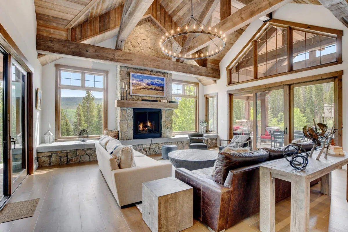 A spacious great room featuring a large stone fireplace, a stylish chandelier, comfortable seating, and expansive windows showcasing breathtaking mountain views. The room’s wooden beams and warm lighting add rustic charm, while the cozy seating arrangement invites guests to relax and enjoy the scenery or gather around the fire.